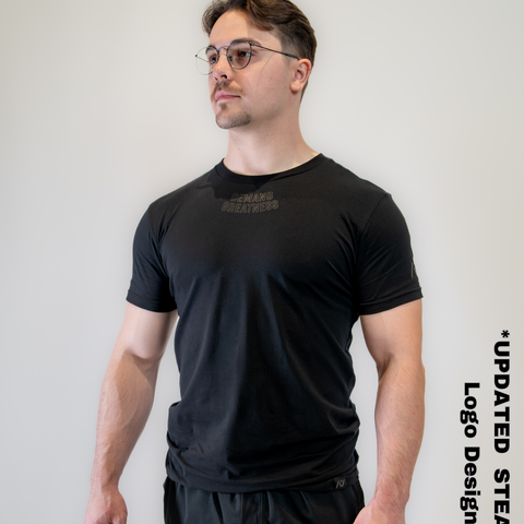 Man wearing A7's IPF Approved Stealth Meet shirt in black. 'Demand Greatness’ written on the front in a dark grey double outlined font to have impact on the competition platform. Paired with black shorts made from a stretchy squat proof material to use in powerlifting training.
