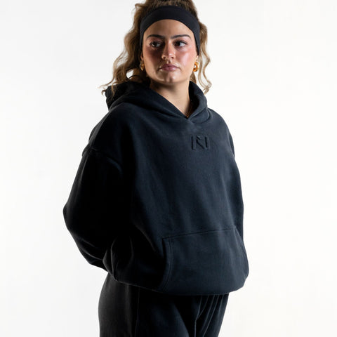 Front of the A7 Veil overfit hoodie in stealth/black. Worn by a woman and unisex sizing, this strength sports apparel is meet day apparel for traveling to meets and standing on the podium 