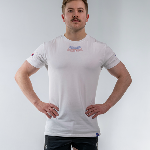 Man wearing A7's IPF Approved white GBR Meet shirt. 'Demand Greatness’ written on the front in a red and blue double outlined font to have impact on the competition platform. Paired with black shorts made from a stretchy squat proof material to use in powerlifting training.
