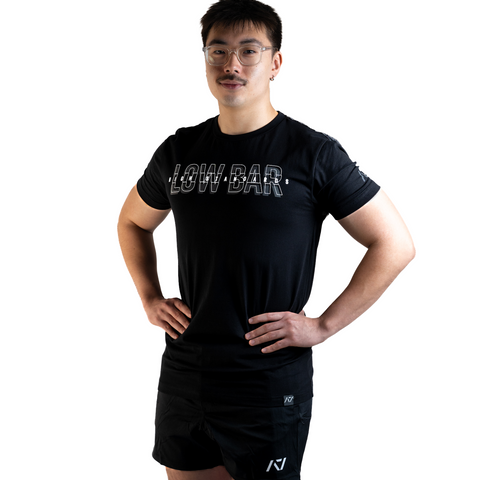 Front View of Man wearing A7's Bar Grip High Standards Low Bar Black shirt. 'HIGH STANDARDS LOW BAR’ written on the front in grey outlined font. Paired with black KWD 360 GO shorts made from a stretchy squat proof material to use in powerlifting training