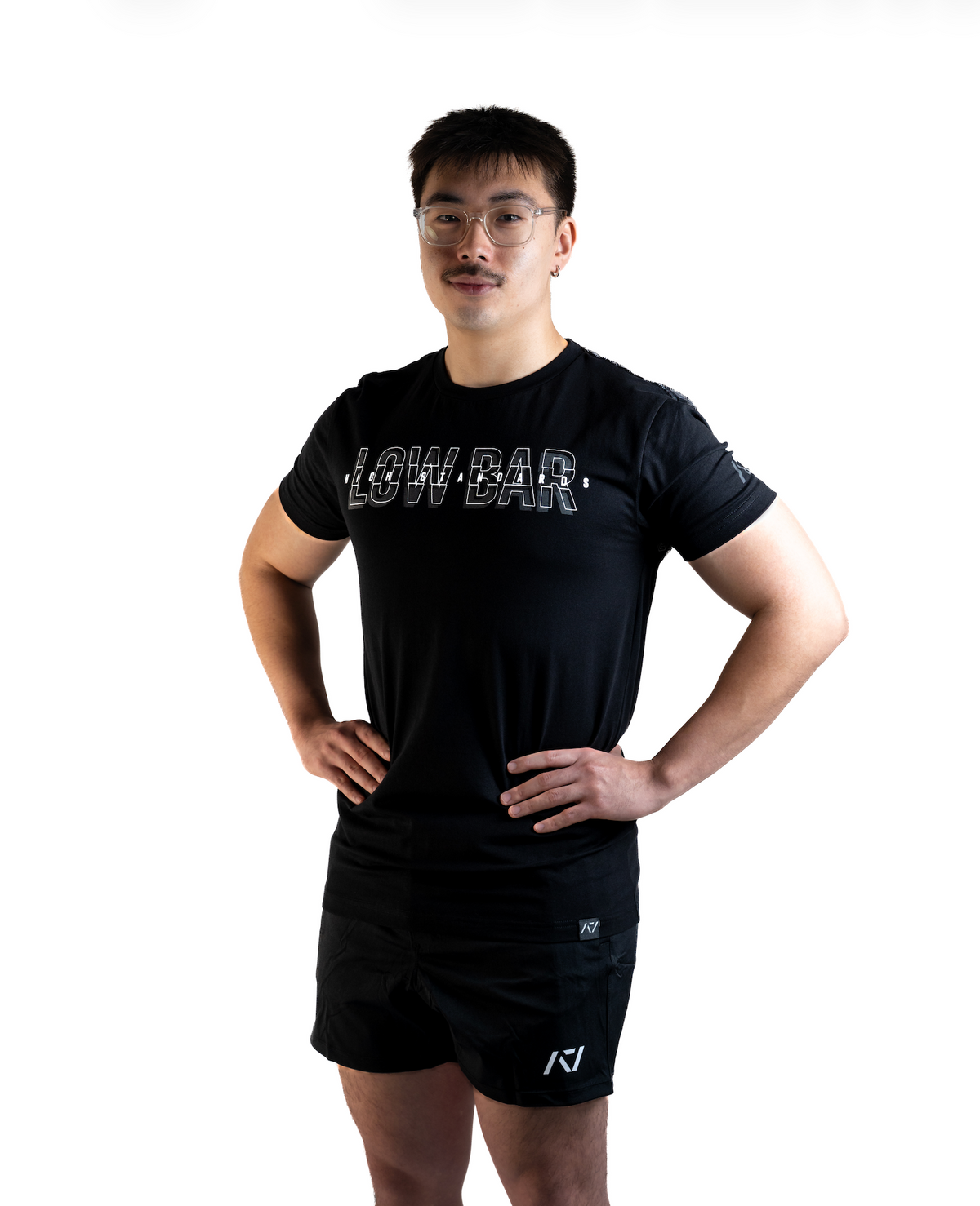 Front View of Man wearing A7's Bar Grip High Standards Low Bar Black shirt. 'HIGH STANDARDS LOW BAR’ written on the front in grey outlined font. Paired with black KWD 360 GO shorts made from a stretchy squat proof material to use in powerlifting training