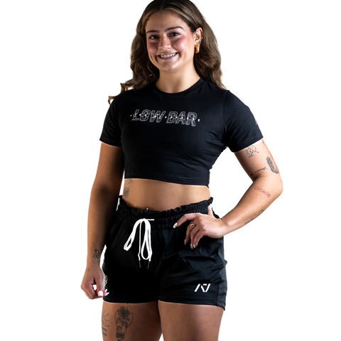 Font View of Woman wearing A7's Bar Grip High Standards Low Bar Black shirt. 'HIGH STANDARDS LOW BAR’ written on the front in grey outlined font. Paired with black KWD 360 GO shorts made from a stretchy squat proof material to use in powerlifting training