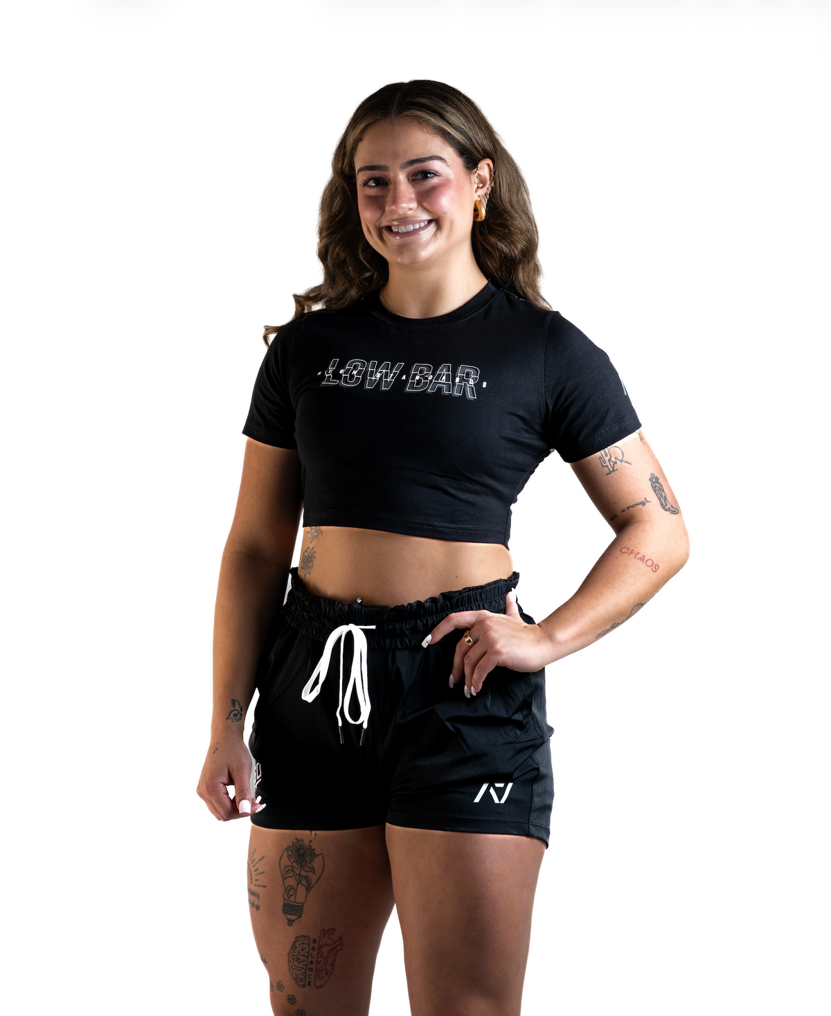 Font View of Woman wearing A7's Bar Grip High Standards Low Bar Black shirt. 'HIGH STANDARDS LOW BAR’ written on the front in grey outlined font. Paired with black KWD 360 GO shorts made from a stretchy squat proof material to use in powerlifting training