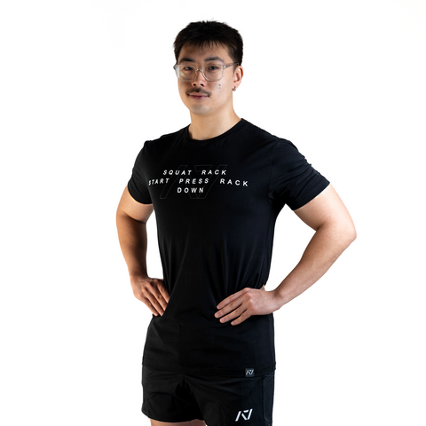 Front View of Man wearing A7's Bar Grip COMMANDS shirt. The Powerlifting Commands for Squat, Bench and Deadlift, written on the front in white and A7 logo outlined behind. Paired with black KWD 360 GO shorts made from a stretchy squat proof material to use in powerlifting training