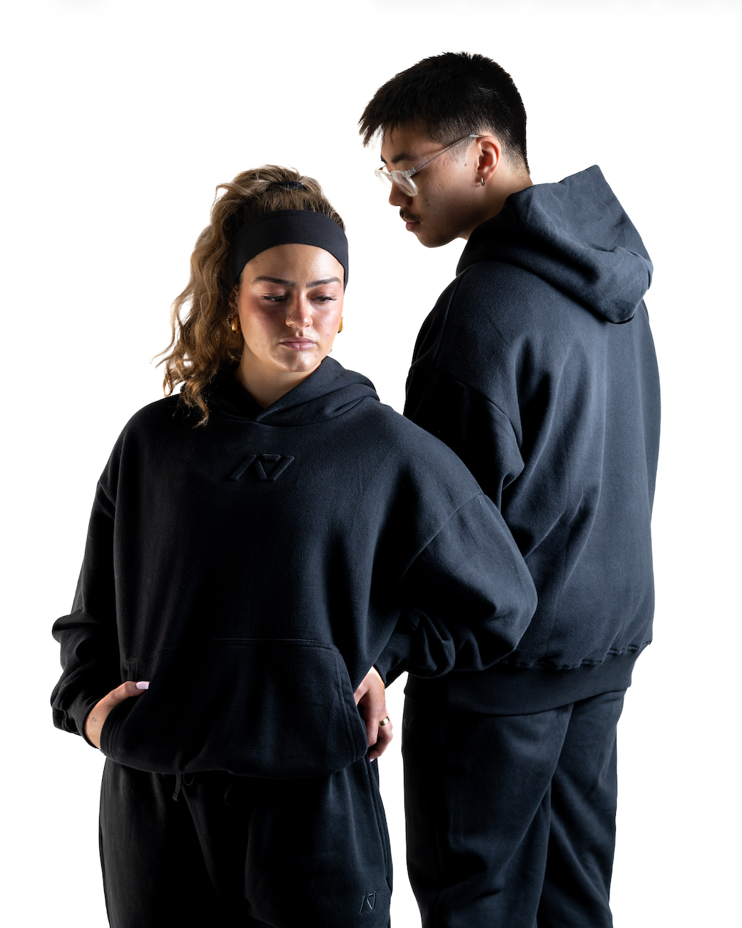 Dual shot of the A7 Veil overfit hoodie in stealth/black. Worn by a man and women showing the front of the womens hoodie and back view of the mans. This is unisex sizing, and strength sports apparel is meet day apparel for traveling to meets and standing on the podium 